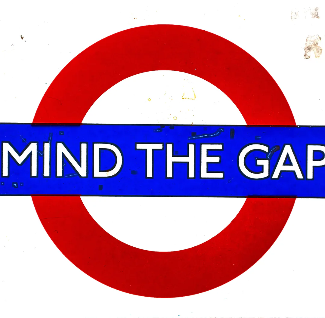 A circular sign with a red outer ring and a blue horizontal bar in the center. The words "MIND THE GAP" are written in large white letters on the blue bar. This sign is commonly found in train stations to remind people to be careful of the space between the train and the platform.